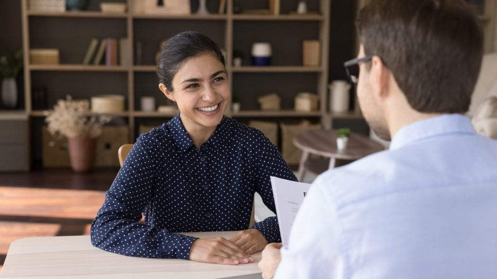 Happy attractive millennial Indian female job seeker holding interview with male leader, making good first impression at meeting, discussing working offer in modern office, employment concept.