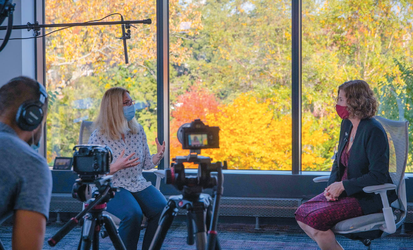 Lisa Pearlman and President Laurie Leshin seated before large windows with fall foliage visible; cameras and a cameraman are visible in the foreground