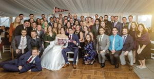a large group of WPI alumni crowd together; a WPI pennant is seen at rear; Juliana (Wakeman) Boucher, in her wedding gown, and Mike Boucher are front and center