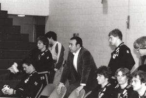 in a black and white photo, Frank DeFalco, in a suit jacket, is with a group of male students in wrestling jackets watching something off camera to the left