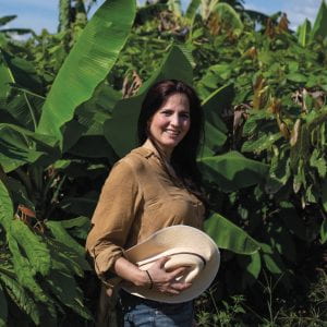 Leila Carvajal Erker, in a tan shirt and holding a white brimmed hat, stands before a field of cacao plants