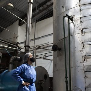 Leila Carvajal Erker, in blue coveralls and a black face mask, stands before a tall silver tank in a factory