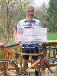 Joel Loitherstein in a decorated t-shirt and blue shorts stands behind a bicycle and holds a large check