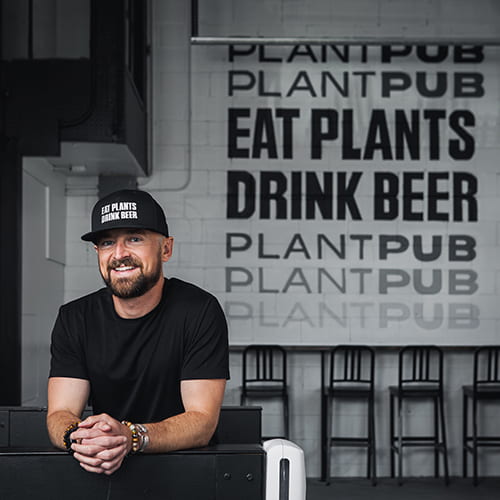 Patrick McAuley in front of a wall mural in his new PlantPub Fenway that reads: Eat Plants Drink Beer